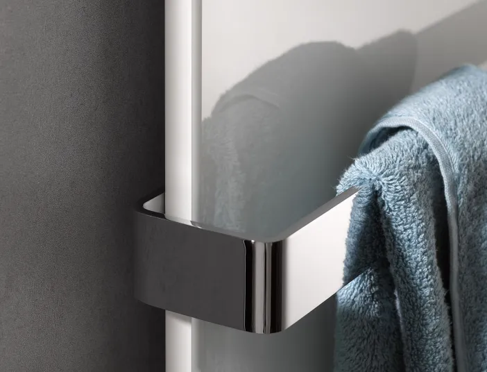 Detailaufnahme eines Glasheizkörpers mit verchromter Halterung, an der ein blaues Handtuch hängt, vor einer dunklen Wand.